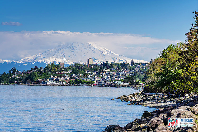 A stunning view of Mount Rainier towering over the cityscape of Tacoma, WA, with the serene waters of Puget Sound in the foreground. The image captures the natural beauty and vibrant community that make living in Tacoma WA appealing. The Mosaic Real Estate logo is visible, showcasing the expertise of Ruthy Taylor, a dedicated Tacoma WA Realtor.